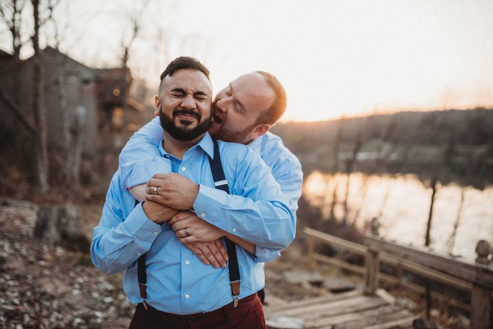 One groom playfully pretends to bite other groom's ear in front of lake